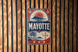 A vintage-style metal sign with the word "MAYOTTE" prominently displayed, featuring a sun and palm tree design.