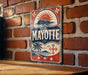 A vintage-style metal sign with the word "MAYOTTE" prominently displayed, featuring a sun and palm tree design, is mounted on a brick wall.