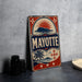 A vintage-style metal sign with the word "MAYOTTE" prominently displayed, featuring a sun and palm tree design. The sign is placed on a gray surface, with some black camera lenses visible in the background.