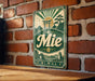 A vintage metal sign with a green and white color scheme, featuring a Japanese-style torii gate and a scenic landscape, mounted on a brick wall.
