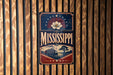 A vintage-style metal sign with the word "MISSISSIPPI" prominently displayed, featuring a riverboat illustration and a flower design.