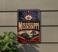A vintage-style metal sign with the word "MISSISSIPPI" prominently displayed, featuring a large white flower and a river scene.