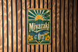 A vintage-style metal sign with the text "Miyazaki" displayed prominently in the center, surrounded by tropical elements such as palm trees and a sun.