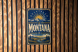 A vintage-style metal sign with the word "Montana" prominently displayed, featuring a mountain landscape and sunburst design.