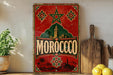 a vintage Moroccan-themed sign with a red background, green and gold accents, and a star at the top. The sign displays the word "MOROCCO" in large white letters. The sign is placed on a wooden surface, with a potted plant to the left, a wooden cutting board to the right, and a bunch of lemons in the foreground.