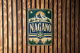 A vintage metal sign with the word "NAGANO" displayed prominently in the center, surrounded by a mountain landscape and snowflake design.