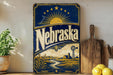A vintage-style metal sign with the word "Nebraska" prominently displayed, featuring a sun and windmill illustration, is placed on a wooden surface next to a potted plant and a wooden cutting board.