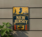 A vintage-style metal sign with the text "NEW JERSEY" prominently displayed, featuring a lighthouse, ferris wheel, and other nautical elements.