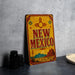 A vintage-style sign for New Mexico, featuring a desert landscape and sun motif, is displayed on a countertop next to some black camera lenses.