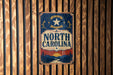 A vintage-style metal sign with the text "NORTH CAROLINA" displayed prominently in the center, surrounded by a decorative border.
