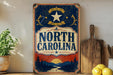 A vintage-style sign for North Carolina, featuring a sun and mountain landscape, is displayed on a shelf next to a wooden cutting board and some lemons.