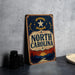 A vintage-style metal sign with the text "NORTH CAROLINA" prominently displayed, standing on a gray countertop next to a stack of black camera lenses.