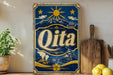 A vintage-style metal sign with the word "Qita" prominently displayed, featuring a sun, mountains, and other scenic elements. The sign is placed on a wooden surface, with a potted plant, a wooden cutting board, and some lemons nearby.