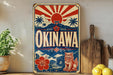 A vintage-style metal sign with the text "OKINAWA" prominently displayed, featuring a red and white sunburst design, a Japanese-style torii gate, and a red and white lion. The sign is placed on a wooden surface, with a potted plant, a wooden cutting board, and some lemons nearby.