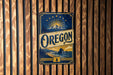 A vintage-style metal sign with the word "Oregon" prominently displayed, featuring a mountain landscape and sun in the background.