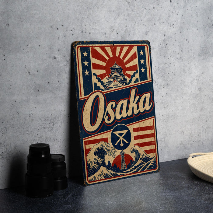 A vintage-style metal sign with the word "Osaka" prominently displayed, featuring a wave and a castle-like structure. The sign is placed on a gray countertop, with some black camera lenses visible to the left.