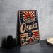 A vintage-style metal sign with the word "Osaka" prominently displayed, featuring a wave and a castle-like structure. The sign is placed on a gray countertop, with some black camera lenses visible to the left.