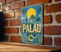 A vintage metal sign with the word "PALAU" displayed prominently, featuring a tropical island landscape with palm trees and a large yellow sun in the sky.