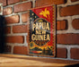 A vintage-style metal sign with the text "PAPUA NEW GUINEA" displayed prominently, featuring a tropical island landscape and a bird in flight.