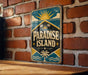 A vintage-style sign for "Paradise Island" is displayed on a brick wall, featuring a sun, palm trees, and a tropical landscape.