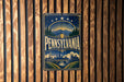 A vintage-style metal sign with the word "PENNSYLVANIA" prominently displayed, featuring a scenic illustration of the state's landscape and architecture.