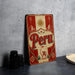 a vintage-style metal sign with the word "Peru" prominently displayed, along with a decorative border and a sun motif. The sign is placed on a gray countertop, and there are some black camera lenses visible in the background.