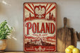 a vintage-style metal sign with the word "POLAND" prominently displayed, surrounded by a red and white design with a castle and mountains. The sign is placed on a wooden surface, with a potted plant, a wooden cutting board, and some lemons nearby.