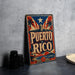 A vintage-style metal sign with the text "PUERTO RICO" prominently displayed, featuring a red, white, and blue color scheme with a star and palm trees. The sign is placed on a gray surface, with a stack of black camera lenses and a white dish visible in the background.
