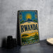 A vintage-style metal sign with the word "Rwanda" prominently displayed, featuring a sun and mountain landscape design.