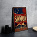 A vintage-style metal sign with the word "SAMOA" prominently displayed, featuring a red, blue, and gold color scheme with a tropical island and sun motif.