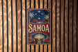 A vintage-style sign with the word "SAMOA" prominently displayed, featuring a tropical landscape with palm trees, mountains, and a sun.