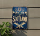 A metal sign with the word "SCOTLAND" prominently displayed, featuring a shield, a lion, and a deer. The sign is mounted on a wall, and there is a plant to the left of the sign.