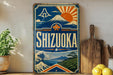 A vintage-style sign for Shizuka, a Japanese beverage, featuring a mountain, sun, and waves, along with the text "SHIZUOKA" prominently displayed.