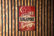 A vintage-style sign for Singapore, featuring a crescent moon and stars, a city skyline, and a lion, is displayed on a wooden wall.