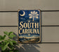 A vintage-style metal sign with the text "SOUTH CAROLINA" displayed prominently, featuring a palm tree and other coastal elements.