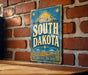 A vintage-style sign for South Dakota is displayed on a brick wall, featuring a sun and mountain landscape design.