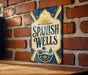 A vintage-style sign for "Spanish Wells" is displayed on a brick wall, featuring a sun and palm tree design.