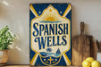 A vintage-style sign for "Spanish Wells" is displayed on a shelf, accompanied by a potted plant, a cutting board, and some lemons.