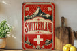 a vintage-style sign for Switzerland, with a mountain and a Swiss cross in the center. The sign is red and white, and it has a vintage appearance. The sign is displayed on a shelf, and there are some kitchen utensils, such as a cutting board and a knife, next to it.