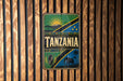 A vintage-style sign with the word "TANZANIA" prominently displayed, featuring a sun, mountain, and other African-inspired elements.