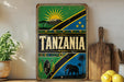 A vintage-style sign with the word "TANZANIA" prominently displayed, featuring a sun, mountain, and other cultural symbols. The sign is placed on a shelf next to a wooden cutting board and a potted plant.