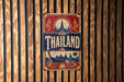 A vintage metal sign with the word "Thailand" prominently displayed, featuring a temple and other traditional Thai imagery.