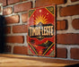 A vintage-style metal sign with a red background and gold accents, featuring a sun and palm trees, is mounted on a brick wall.
