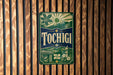 A vintage-style sign with the word "Tochigi" prominently displayed, featuring a sun, mountains, and a bridge.