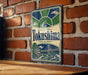 A vintage metal sign with the name "Tokushima" displayed prominently, featuring a bridge and a sun-like symbol. The sign is mounted on a brick wall, and there is a wooden table in the foreground.