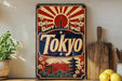 A vintage-style metal sign with the word "Tokyo" prominently displayed, featuring a sunburst design, Japanese-style buildings, and a cherry blossom tree. The sign is placed on a shelf next to a wooden cutting board and a potted plant.
