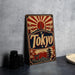 A vintage-style metal sign with the word "Tokyo" prominently displayed, featuring a sunburst design and various Japanese-inspired elements such as pagodas and cherry blossoms.