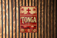 A vintage-style sign with the word "Tonga" prominently displayed, featuring a red cross and palm trees, mounted on a wooden wall.