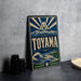A vintage-style sign for Toyama, featuring a mountain landscape and a wave, is displayed on a table next to some black camera lenses.