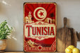 A vintage-style metal sign with the word "TUNISIA" prominently displayed, featuring a red and white color scheme with a crescent moon and star symbol. The sign is placed on a wooden surface, with a potted plant, a wooden cutting board, and some lemons nearby.
